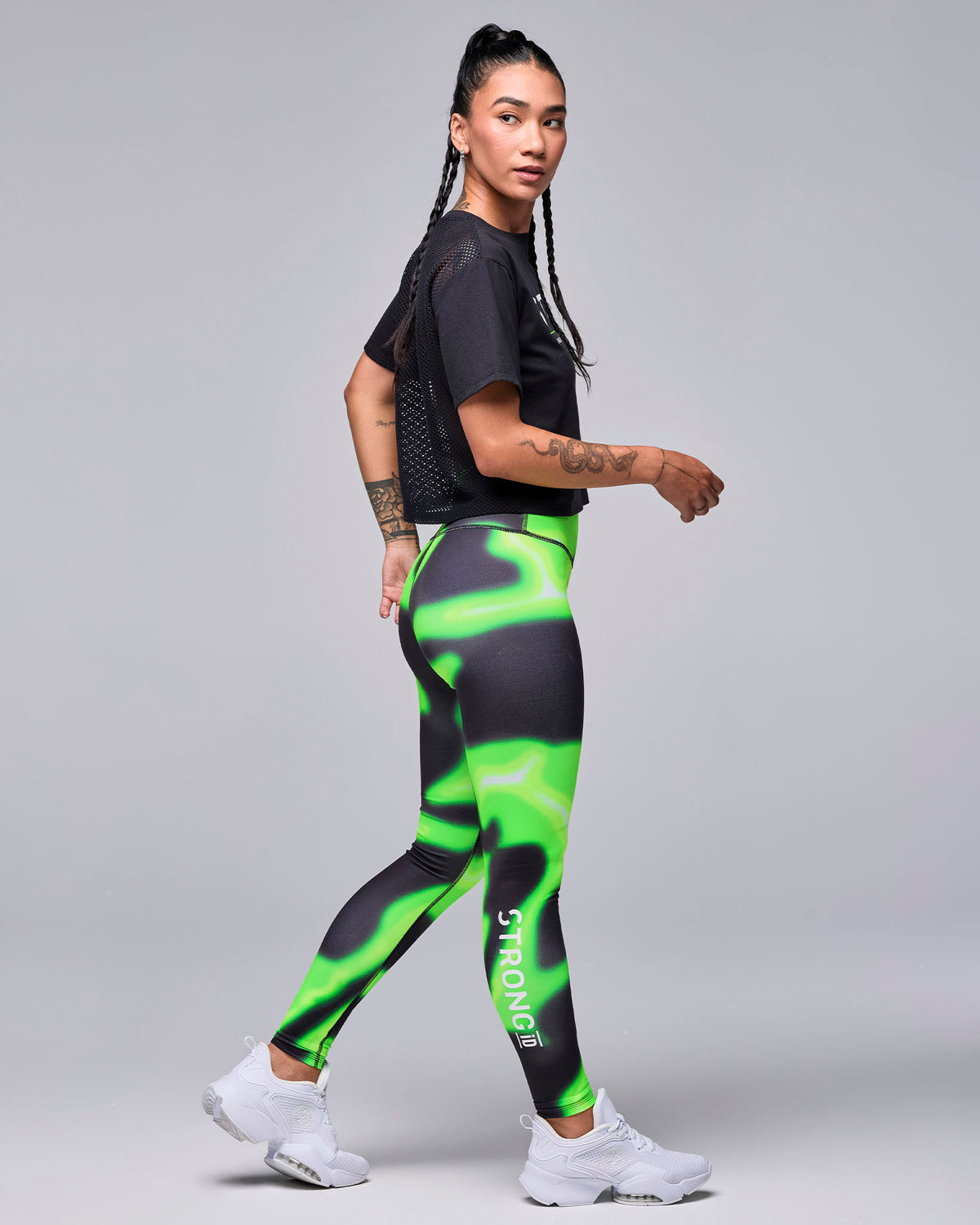 Female model walking in the black 'STRONG' boxy top, shown in side view with the mesh back visible, plus neon green and black swirl print leggings and white shoes.