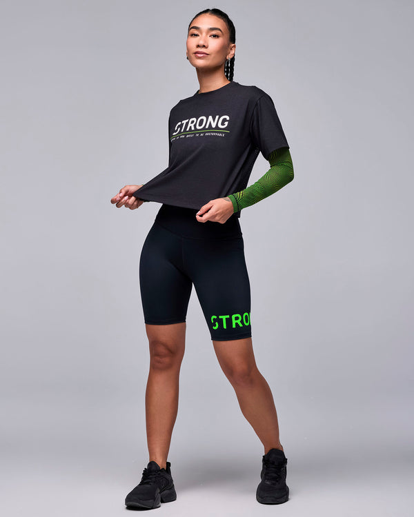 Full view of a female model in the black 'STRONG' boxy top over a wavy check print mesh long sleeve top, with black 'STRONG iD' biker shorts and black shoes.
