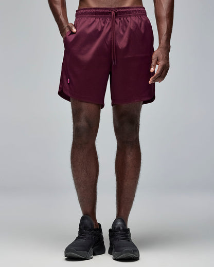 Burgundy athletic shorts with an elastic drawstring waistband, side pocket, curved hem, and small logo tab on the right side. Male model wears the loose-fit shorts in a front angled close-up against a light gray background.