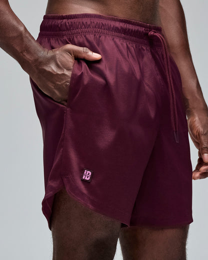 Burgundy athletic shorts with an elastic drawstring waistband, side pocket, curved hem, and small logo tab. Male model wears the loose-fit shorts in a front angled close-up view with his hand in the right pocket showing the small logo and all against a light gray background.