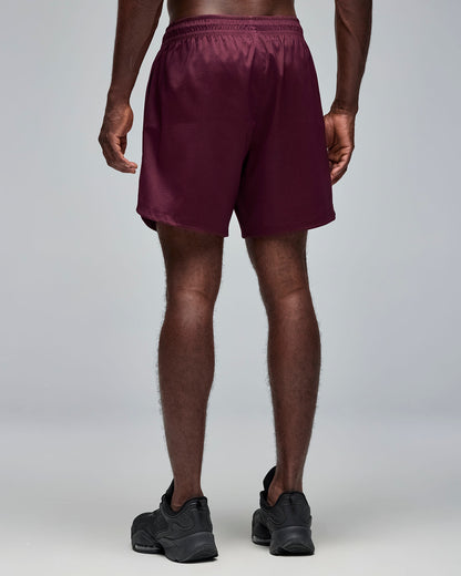 Back view of burgundy athletic shorts with an elastic waistband and relaxed fit. Male model wears the shorts with black athletic sneakers against a light gray background.