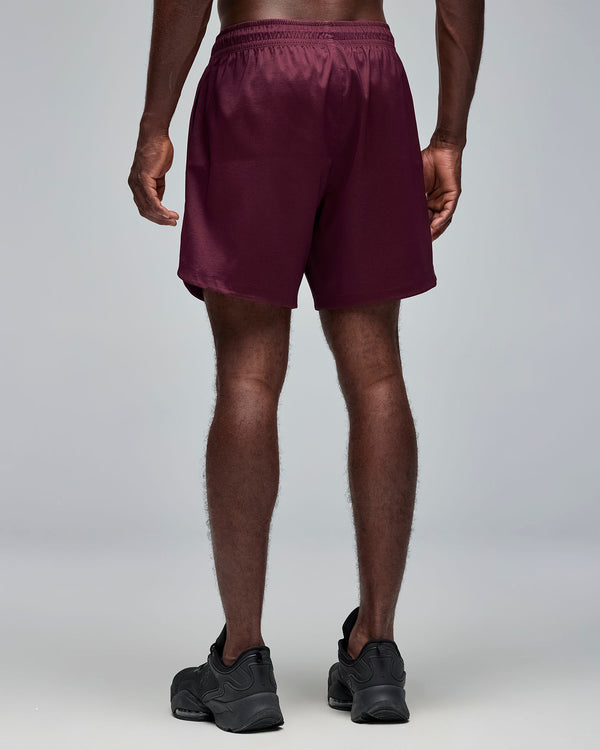 Back view of burgundy athletic shorts with an elastic waistband and relaxed fit. Male model wears the shorts with black athletic sneakers against a light gray background.