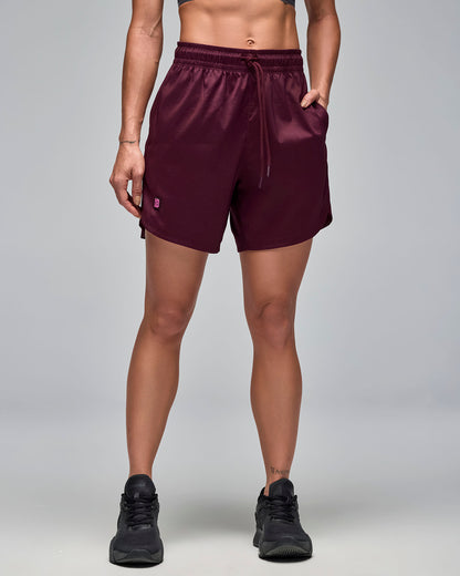 Burgundy athletic shorts with an elastic drawstring waistband and side pockets. Female model wears the loose-fit shorts with black athletic sneakers in a front view against a light gray background with her hand in the left pocket.