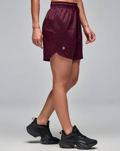 Burgundy athletic shorts with an elastic drawstring waistband, side pocket, and curved hem with a small logo tab. Female model wears the loose-fit shorts with black athletic sneakers in a side view against a light gray background.