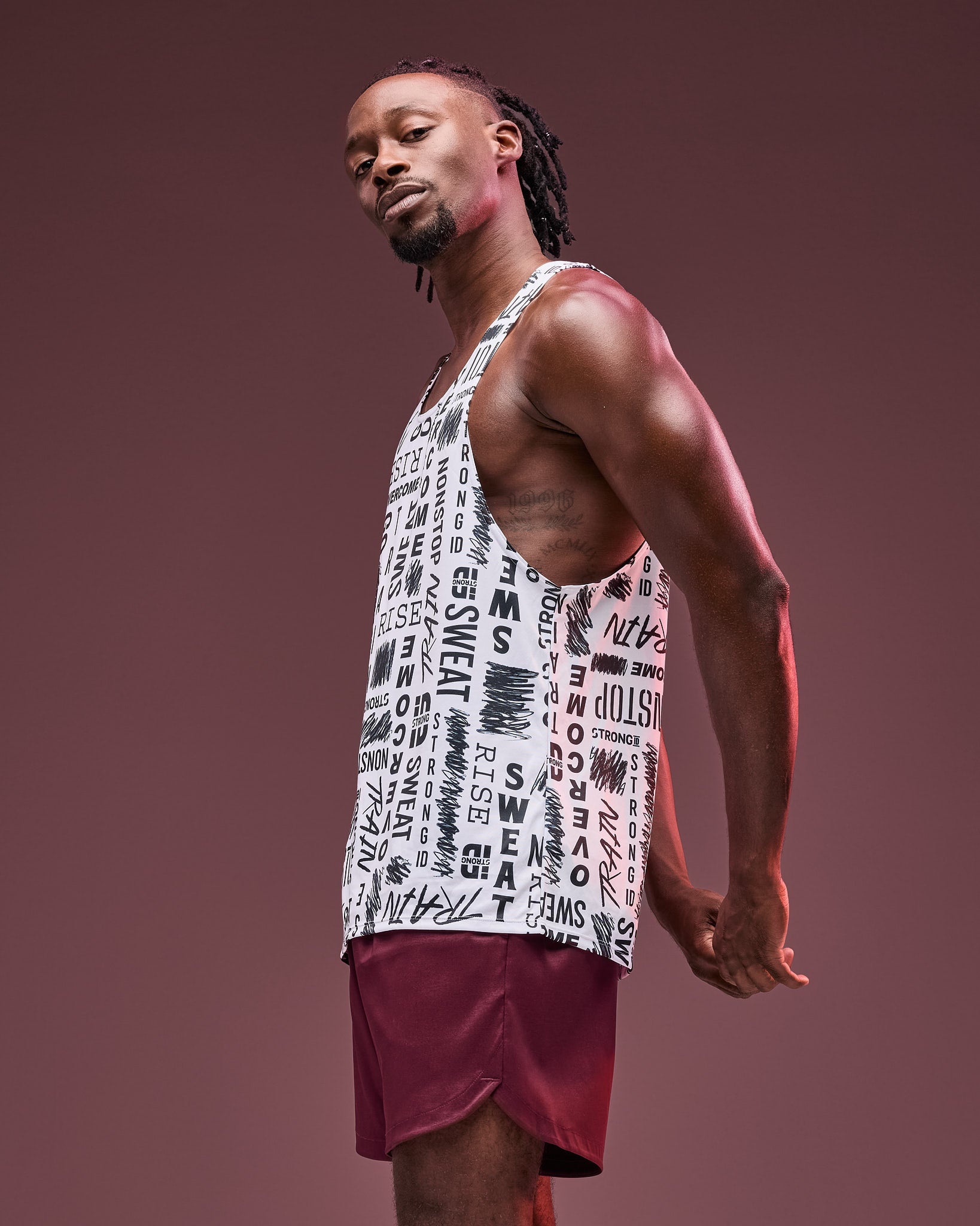 White graphic-print athletic tank with deep-cut armholes, shown in side view on a male model wearing burgundy athletic shorts against a dark mauve studio background.