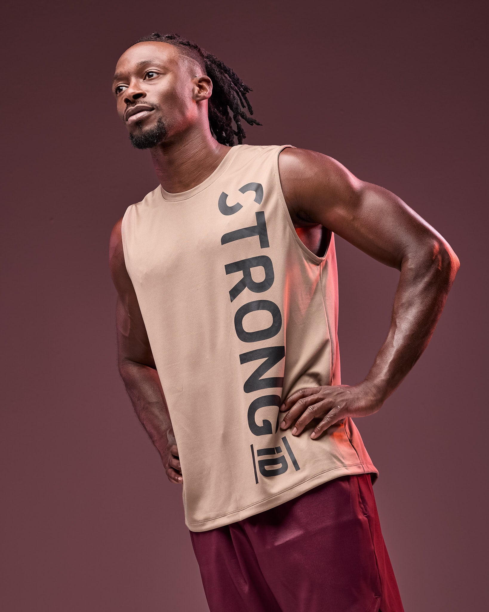 Taupe sleeveless athletic tank with an oversized vertical black "STRONG iD" logo, shown on a male model wearing burgundy athletic shorts against a dark mauve studio background.