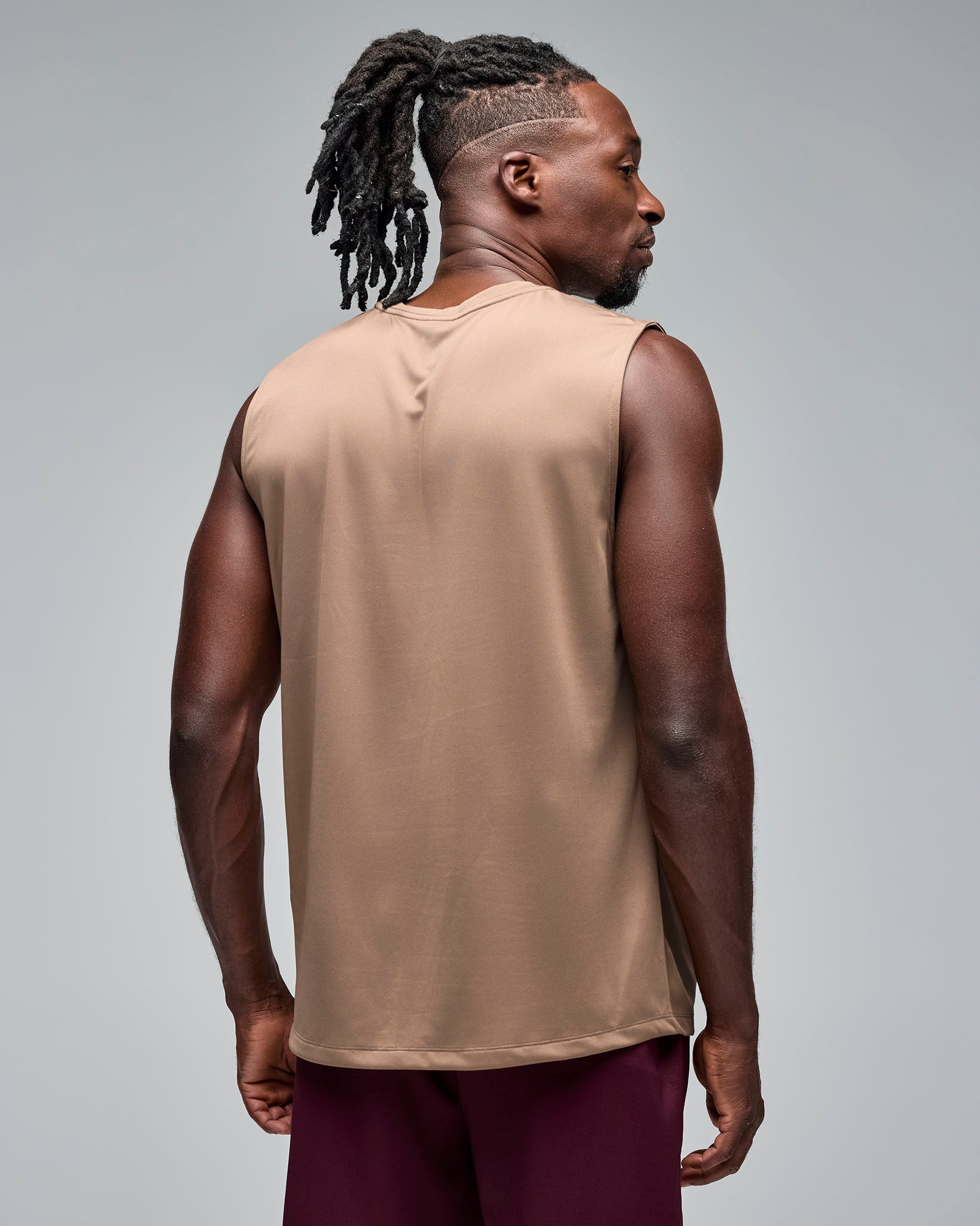 Taupe sleeveless athletic tank with a crew neckline and plain back, shown from the back on a male model wearing burgundy athletic shorts against a light gray studio background.
