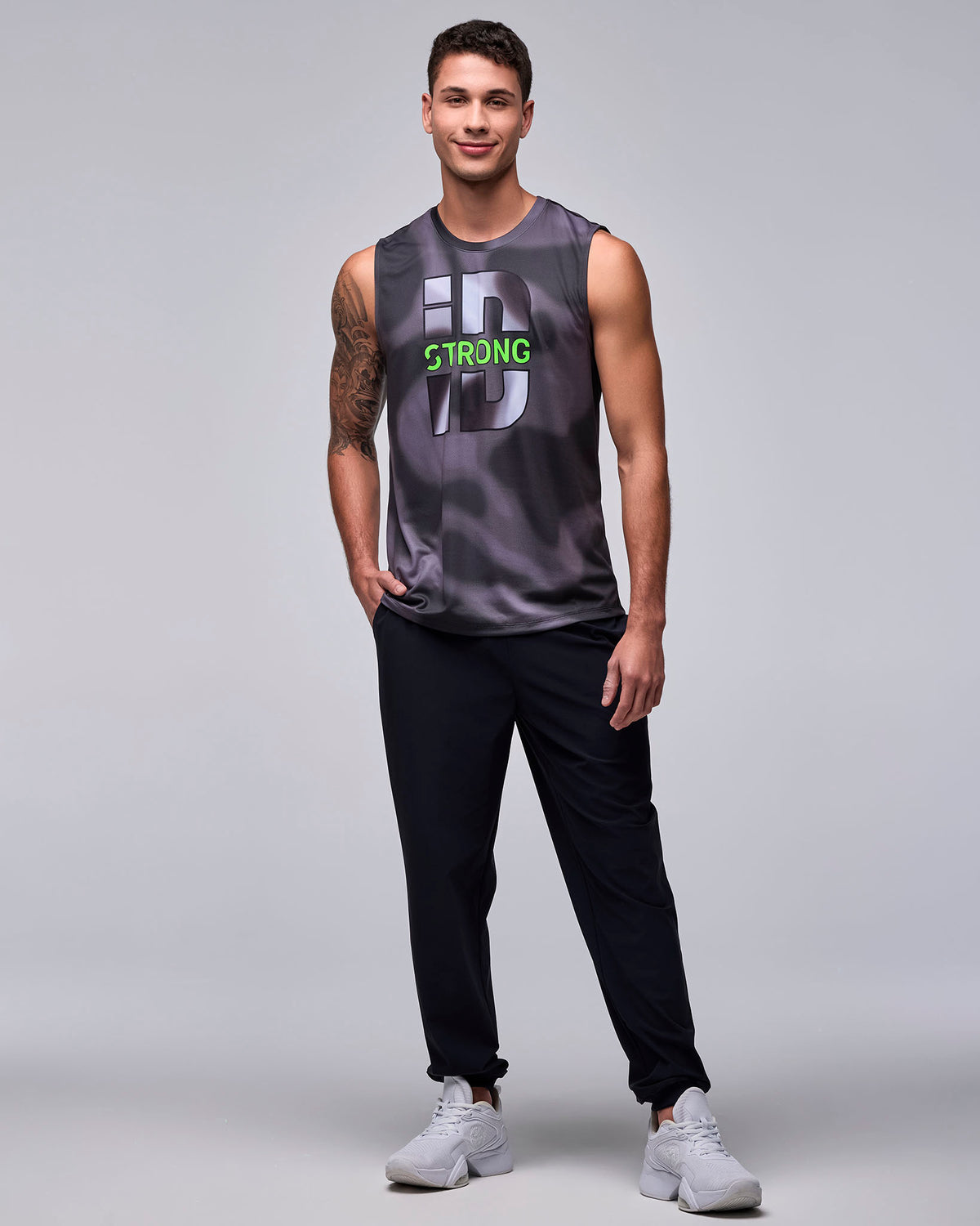 Full view of a male model in the black and grey 'STRONG iD' wide shoulder tank with black joggers and white shoes, one hand in the pocket.