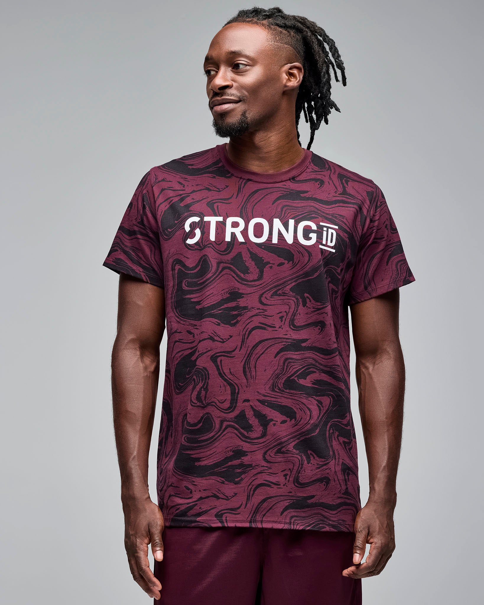 Maroon and black swirl-print graphic T-shirt with bold white “STRONG iD” text on the chest. Male model wears the short-sleeve crewneck tee with burgundy athletic shorts, standing against a light gray background and looking to the side.