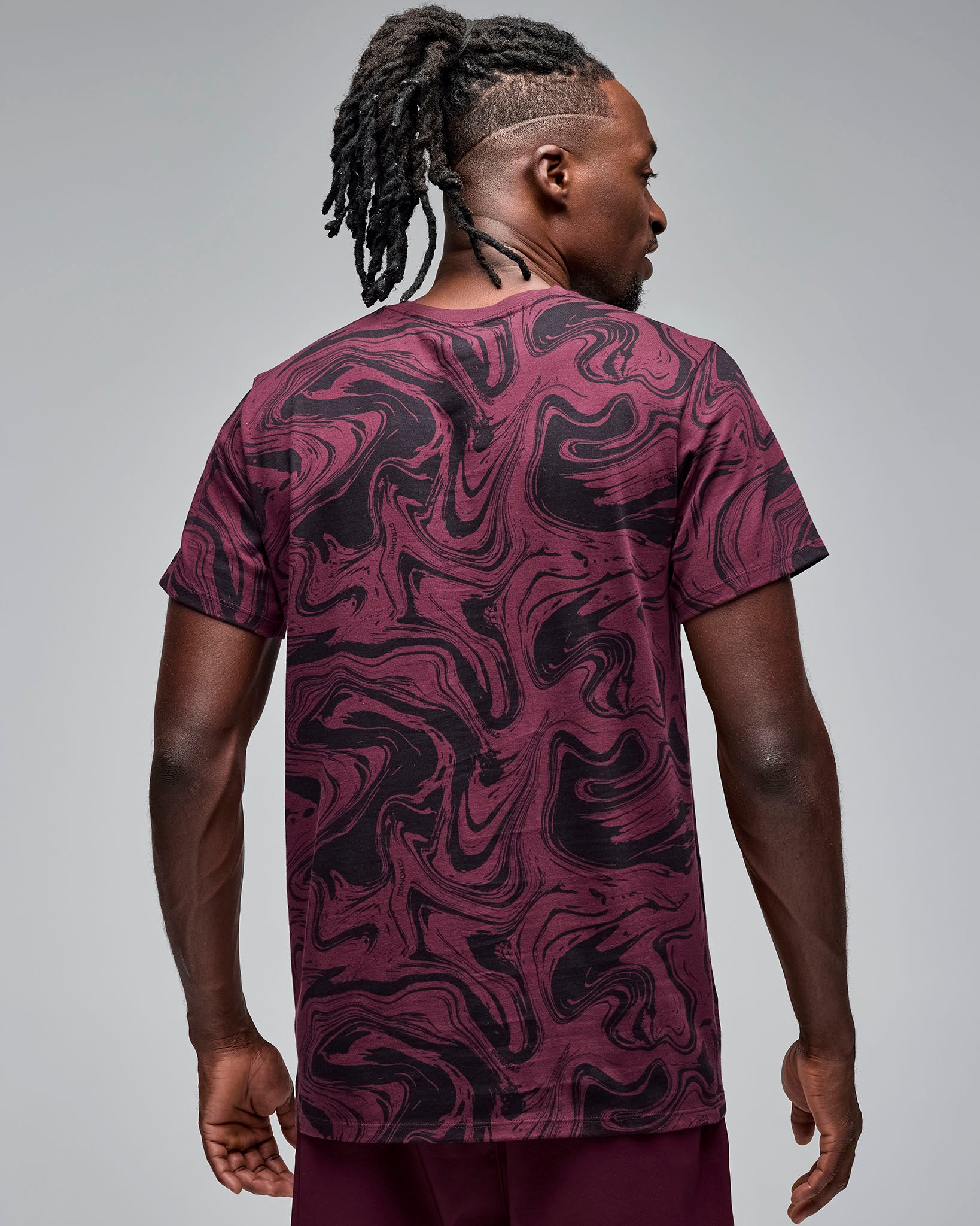 Back view of a maroon and black swirl-print short-sleeve T-shirt. Male model wears the crewneck tee with burgundy athletic shorts, showing the allover abstract pattern on the back against a light gray background.