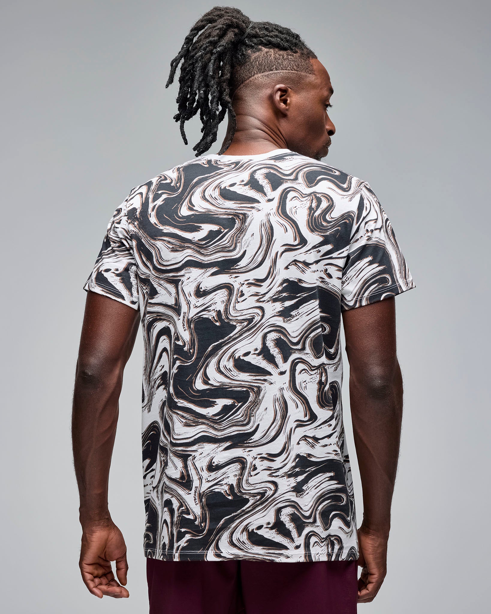 Back view of a white and black swirl-print short-sleeve T-shirt. Male model wears the crewneck tee with burgundy athletic shorts, showing the all-over abstract pattern on the back against a light gray background.