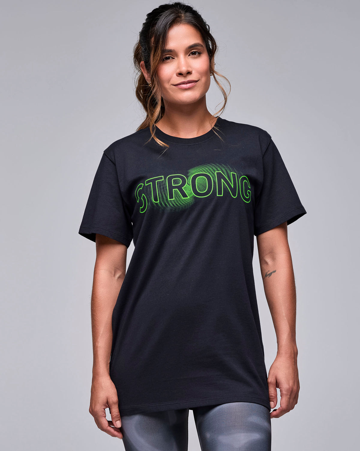 Female model in the black unisex crew neck tee with neon green outlined 'STRONG' text over a green wave-like graphic, worn loose with black and grey swirl print leggings.
