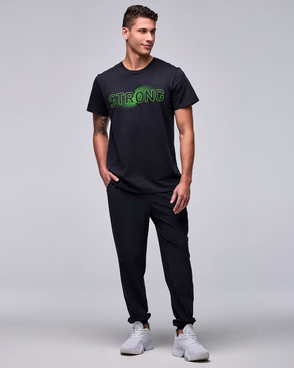 Full view of a male model in the black 'STRONG' wave-like graphic tee with black joggers and white shoes.