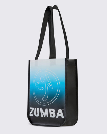 Reusable black tote with black woven handles, turned to show its front panel fading from white at the top through light blue to black, with a large white outline Zumbito and bold white 'ZUMBA' text beside the black side gusset.