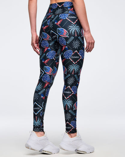 Back view of high-waisted black tropical-print leggings with “ZUMBA” diamond logos and palm-and-parrot graphics; female model wears white sneakers against a white background.