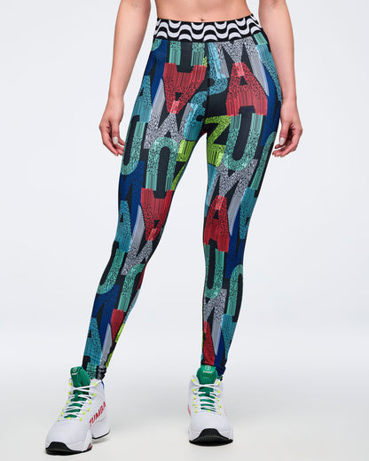 Front view of the multicolor letter-print leggings featuring oversized letter graphics and a black-and-white wavy waistband; female model wears white high-top Zumba sneakers on a white background.