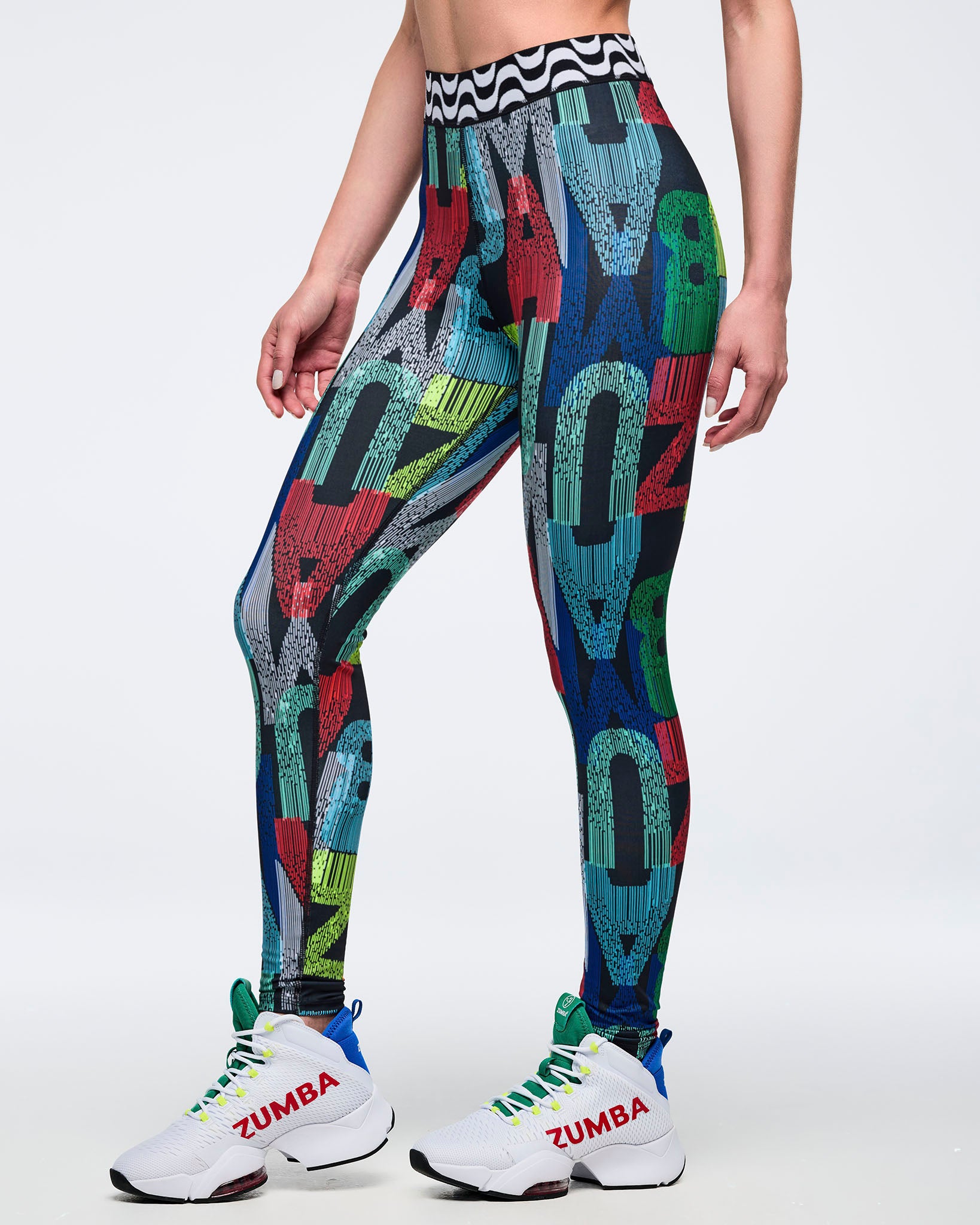 High-waisted multicolor letter-print leggings with a black-and-white wavy waistband; female model wears white high-top Zumba sneakers, shown on a white background.