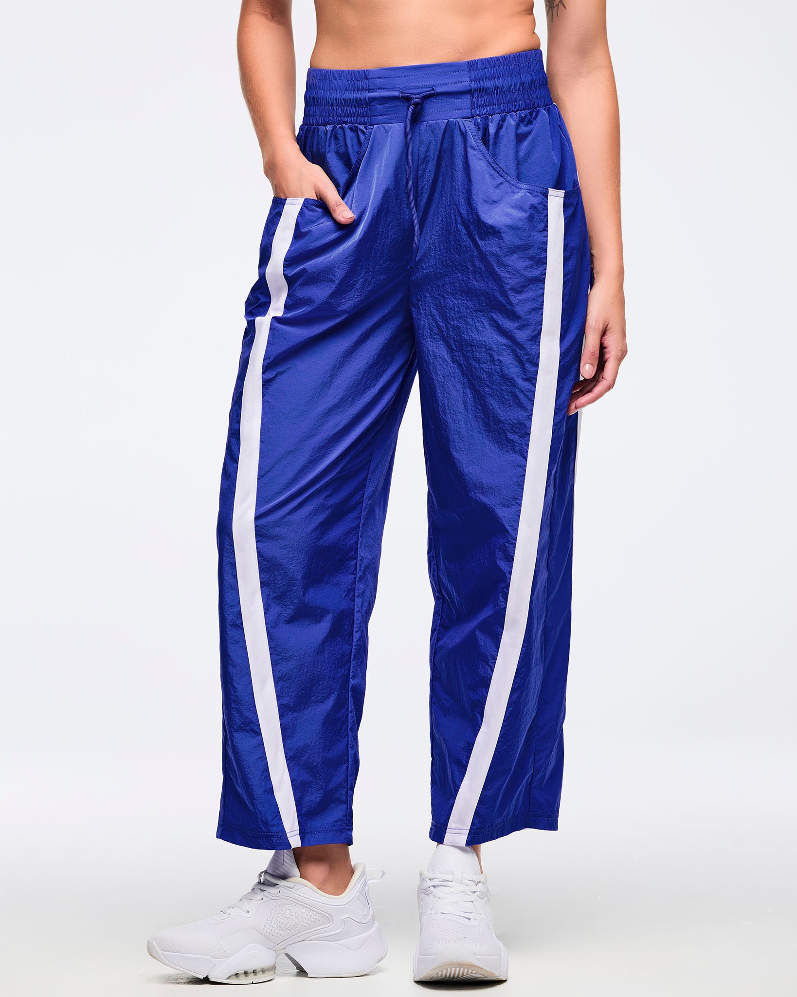 Front view of royal blue wide-leg track pants with white side stripes, elastic drawstring waistband, and front pockets; female model wears white sneakers on a white background.