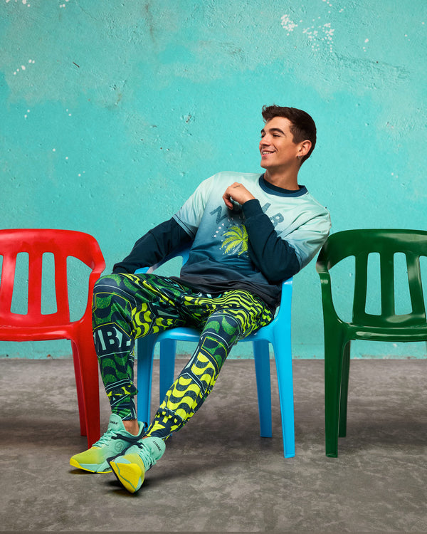 Lifestyle shot of a male model seated on a blue plastic chair between red and green chairs against a turquoise wall, wearing the neon green/teal graphic Zumba jogger pants and teal-and-lime sneakers, with a long-sleeve top.