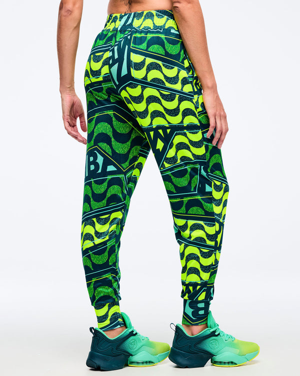 Back view of the neon green/teal graphic Zumba jogger pants with elastic waistband and tapered cuffs; paired with teal-and-lime Zumba sneakers on a white background.