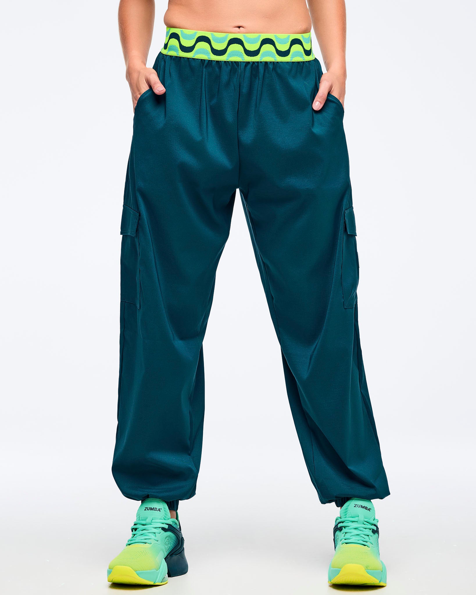 Teal cargo jogger pants with a neon green-and-black wave print elastic waistband, shown from the front on a female model with hands in pockets; loose legs with cargo pockets and elastic cuffs, paired with aqua-to-lime sneakers.