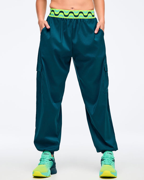Teal cargo jogger pants with a neon green-and-black wave print elastic waistband, shown from the front on a female model with hands in pockets; loose legs with cargo pockets and elastic cuffs, paired with aqua-to-lime sneakers.