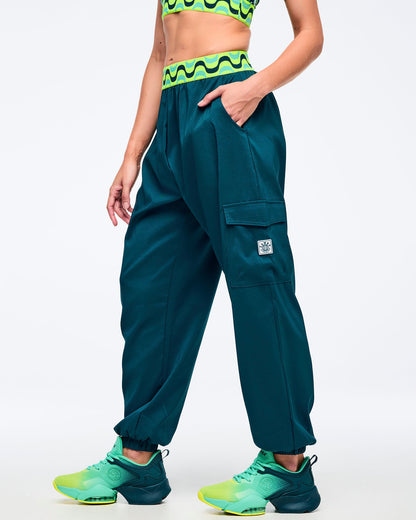Teal cargo jogger pants with a neon green-and-black wave print elastic waistband, shown on a female model in a side pose with hands in pockets; tapered legs with elastic cuffs and aqua-to-lime sneakers.