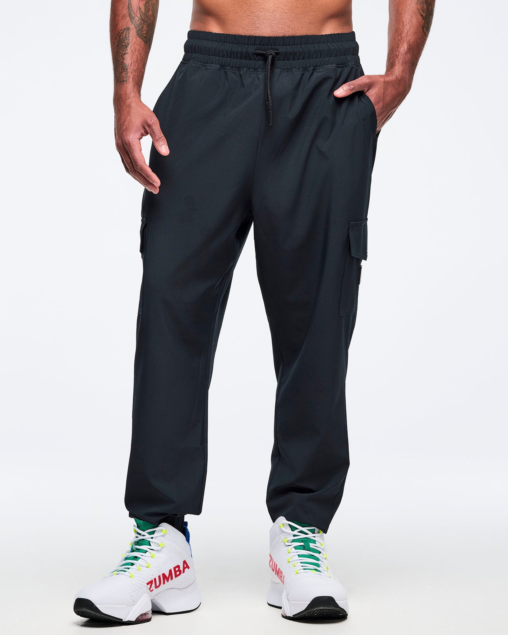 Black cargo jogger pants shown from the front on a male model; elastic drawstring waistband, cargo pocket details, and tapered ankles, paired with white high-top Zumba sneakers.