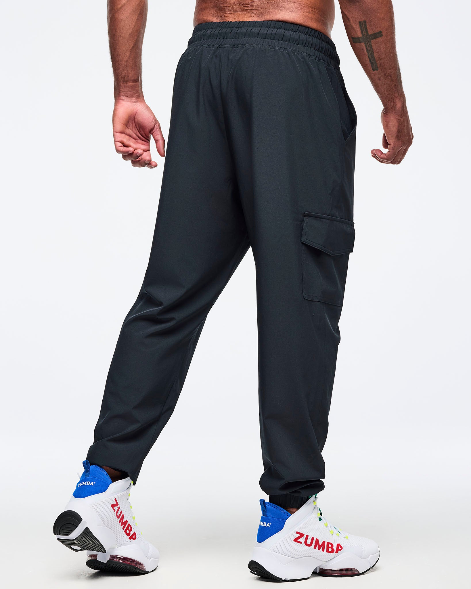 Black cargo jogger pants shown from the back on a male model; relaxed fit with side cargo pocket and elastic waistband, paired with white high-top Zumba sneakers.