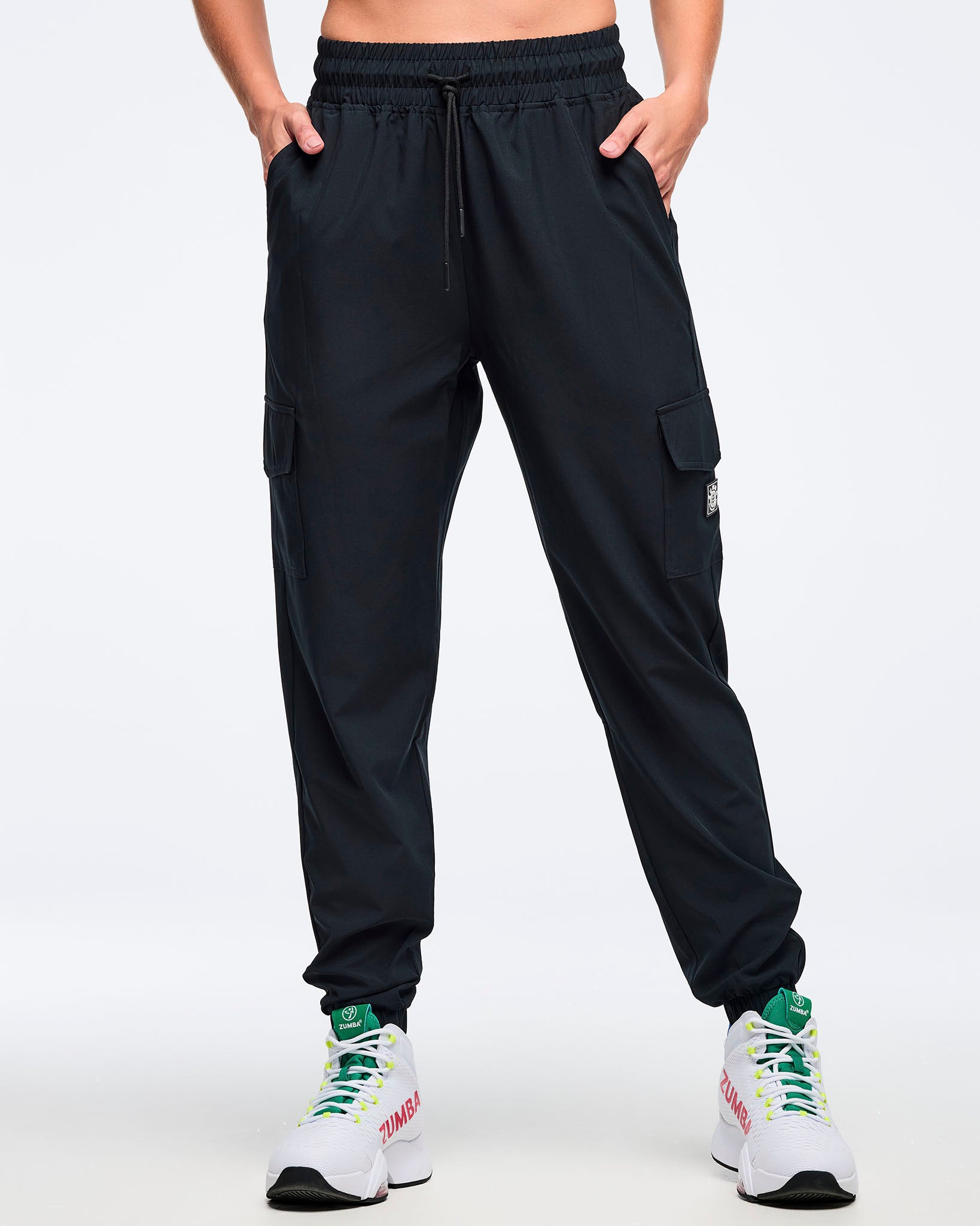Black cargo jogger pants with an elastic drawstring waistband, shown from the front on a female model with hands in pockets; straight-to-tapered legs with cargo pockets and ankle cuffs, paired with white high-top Zumba sneakers.