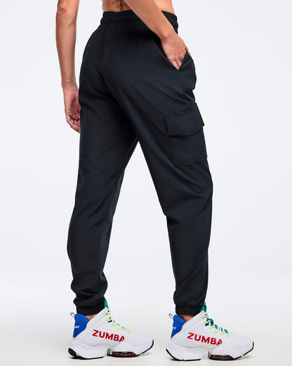 Black cargo jogger pants with an elastic waistband and ankle cuffs, shown from the back on a female model with one hand in a pocket; relaxed fit with side cargo pocket, paired with white high-top Zumba sneakers.