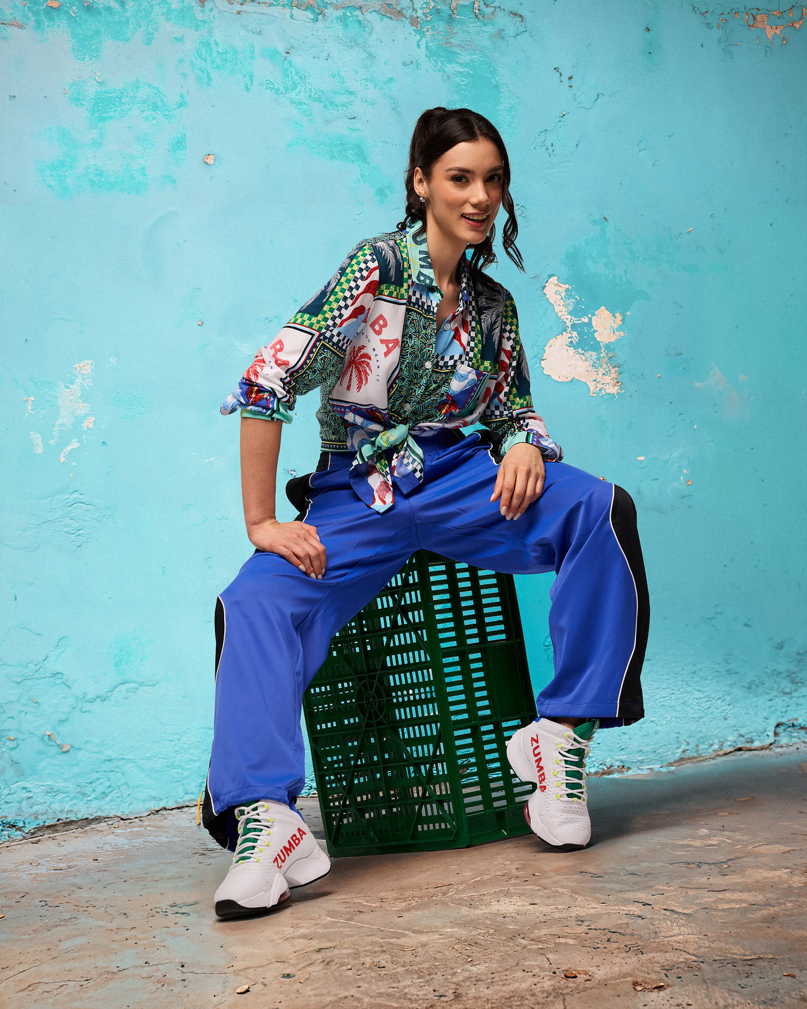 Blue and black track pants with white piping and an elastic waistband, styled on a female model with a colorful printed button-up shirt tied at the waist and white lace-up sneakers, seated on a green crate against a turquoise wall.