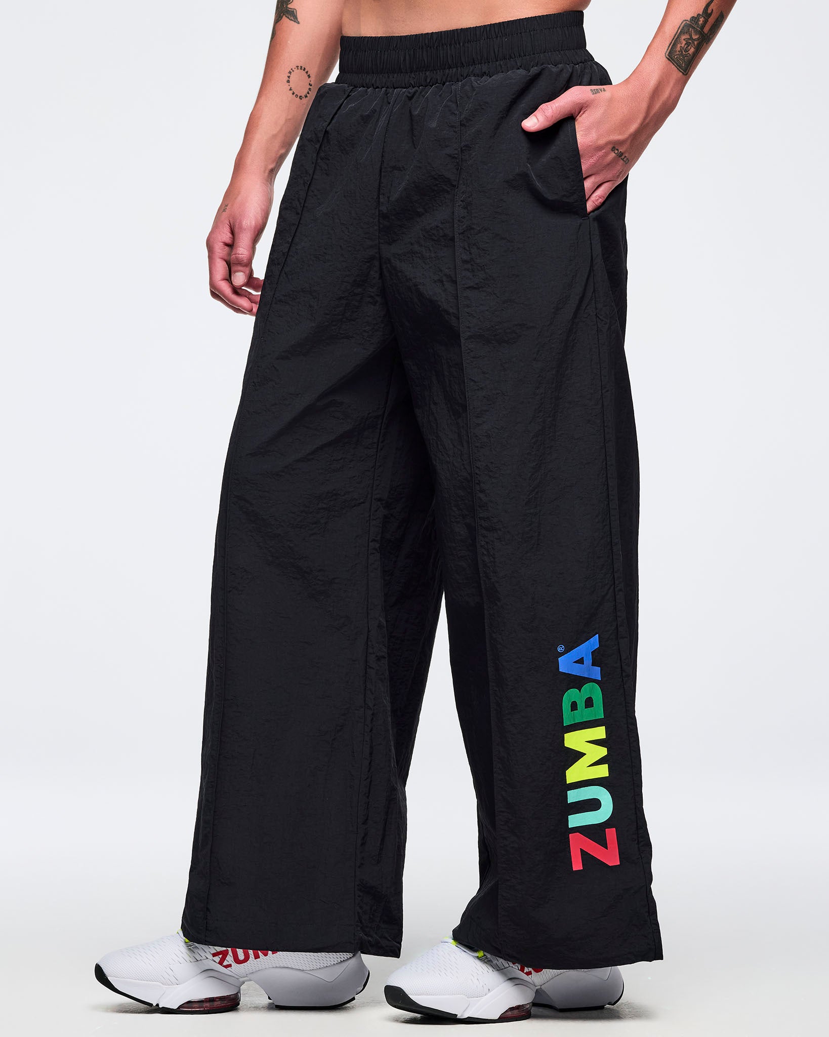 Black wide-leg Zumba pants with elastic waistband and multicolor vertical “ZUMBA” logo near the hem; male model wears white sneakers on a white background.