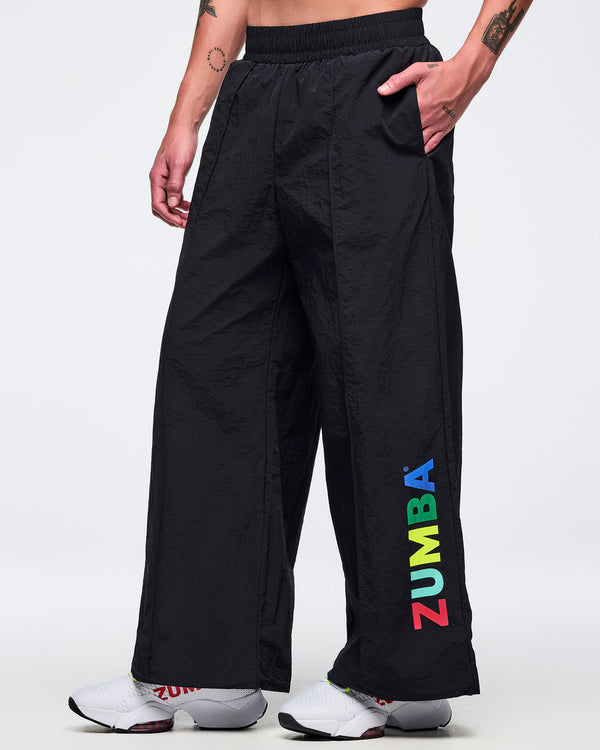 Black wide-leg Zumba pants with elastic waistband and multicolor vertical “ZUMBA” logo near the hem; male model wears white sneakers on a white background.