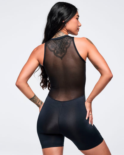 Back view of the bodysuit showing a sheer black mesh racerback-style panel and fitted black biker-short legs, modeled by a female on a white studio background.