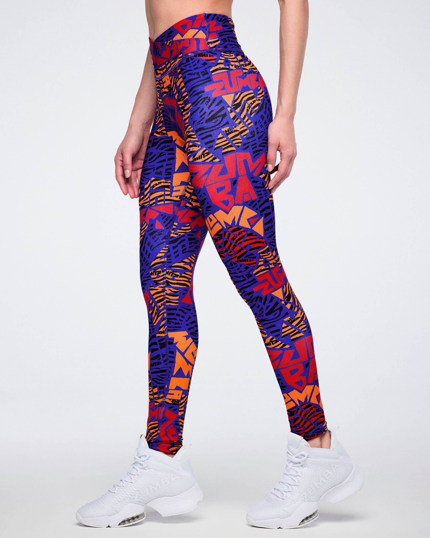 Side view of blue, orange, and red abstract-print high-waisted leggings with a wide crossover-style waistband and fitted full-length silhouette. Female model wears the leggings with white athletic sneakers.