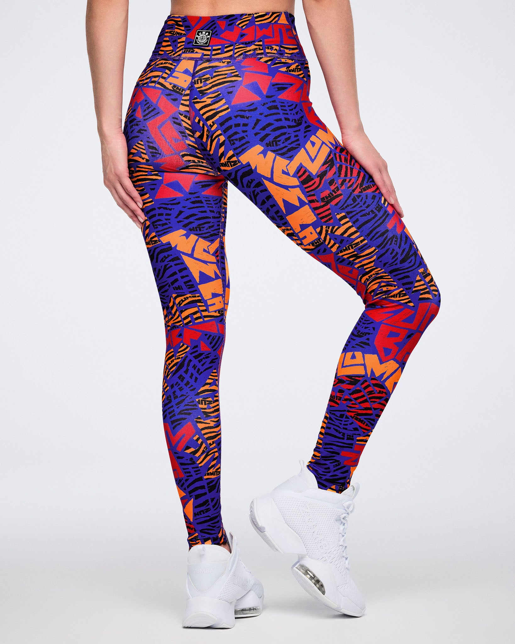 Back view of blue, orange, and red abstract-print high-waisted leggings with a wide waistband, fitted silhouette, and full-length leg. Female model wears the leggings with white athletic sneakers.