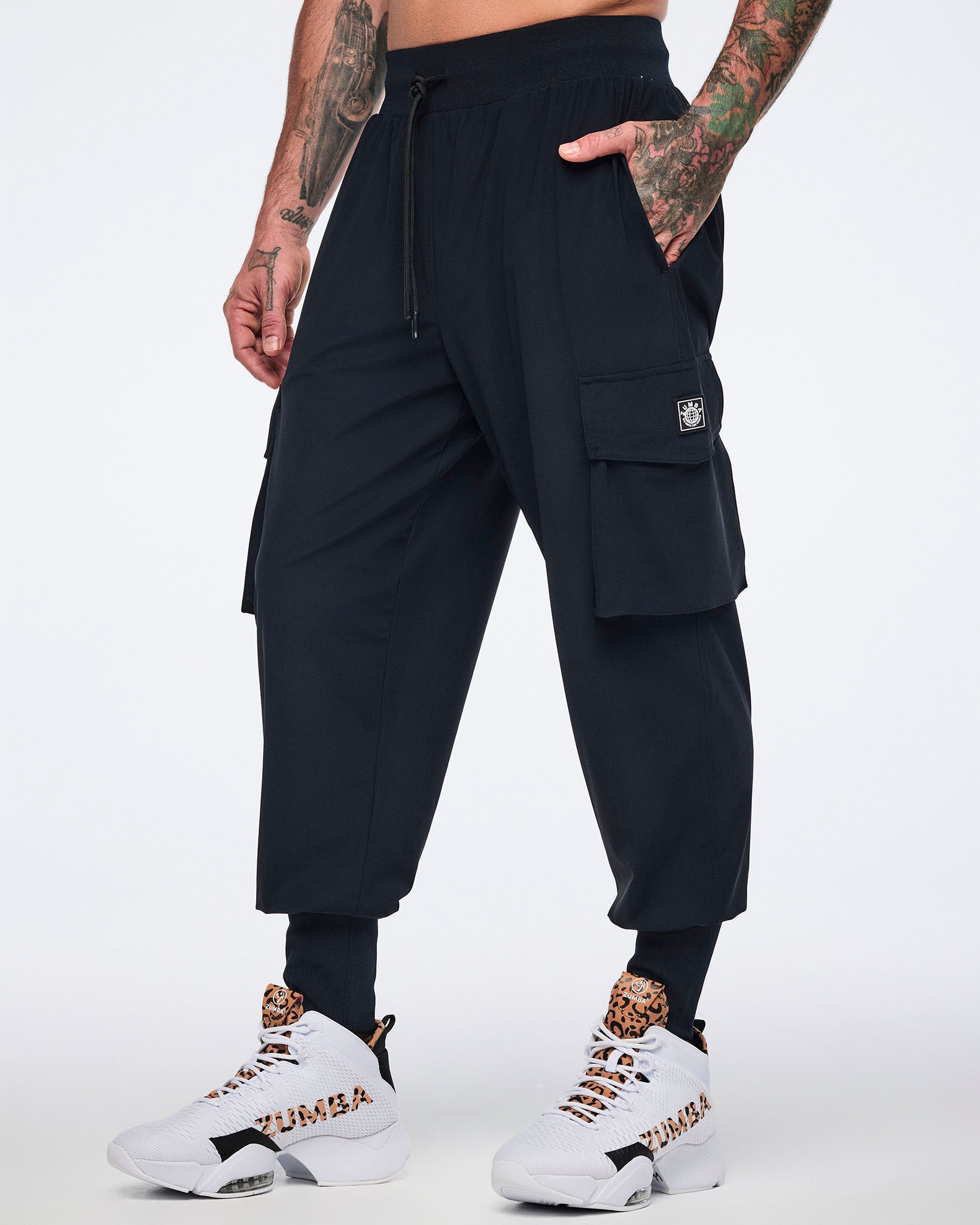 Black cargo jogger pants with oversized flap side pockets, a drawstring waistband, and ribbed ankle cuffs, shown on a male model wearing white high-top sneakers with black and leopard-print details.