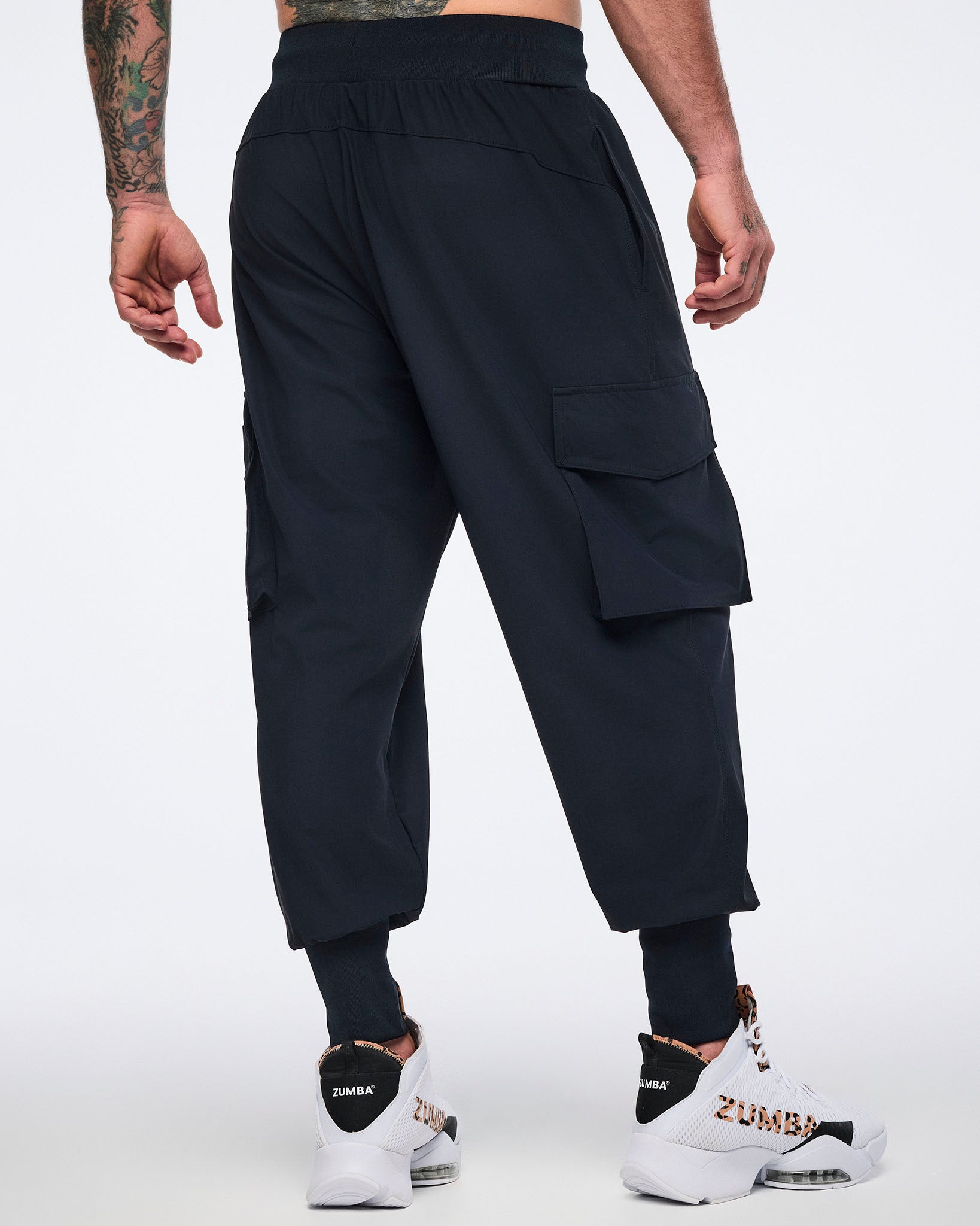 Black cargo jogger pants with oversized flap side pockets, a smooth waistband, and ribbed ankle cuffs, shown from the back on a male model wearing white high-top sneakers with black and leopard-print details.
