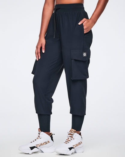 Black cargo jogger pants with oversized flap side pockets, a high-rise waistband with drawstring, and ribbed ankle cuffs, shown from the front on a female model wearing white high-top sneakers with black and leopard-print details.