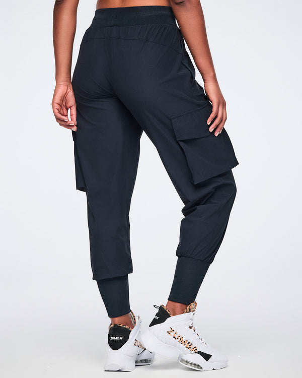 Black cargo jogger pants with oversized flap side pockets, a high-rise waistband with drawstring, and ribbed ankle cuffs, shown from the back on a female model wearing white high-top sneakers with black and leopard-print details.
