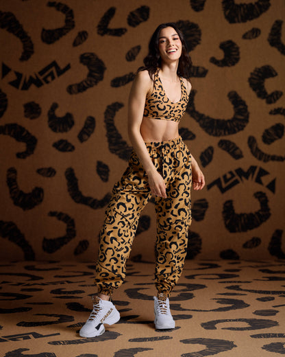 Tan leopard-print cargo pants with black abstract spots and logo accents, shown on a smiling female model in a matching leopard-print sports bra and white high-top sneakers with leopard details, standing against a coordinating leopard-print backdrop.