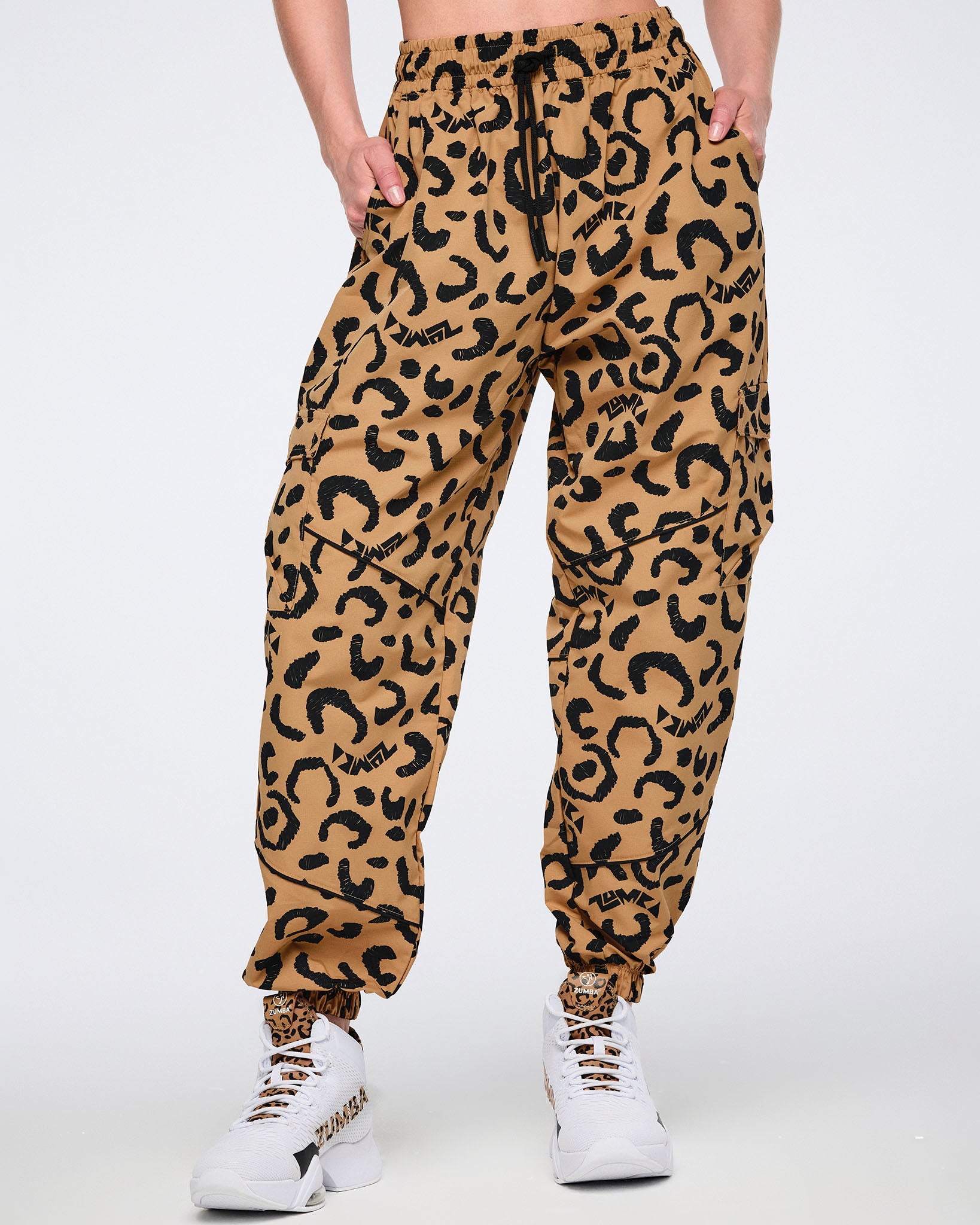Tan leopard-print cargo pants with black abstract spots, a black drawstring waistband, flap side cargo pockets, and elastic cuffs, shown from the front on a female model wearing white high-top Zumba sneakers with black and leopard-print accents.