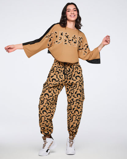 Tan leopard-print cargo pants with black abstract spots and elastic cuffs, shown on a female model wearing a tan oversized cropped top with leopard-print “Zumba” lettering, black mesh sleeve panels, and white high-top sneakers with leopard-print details.