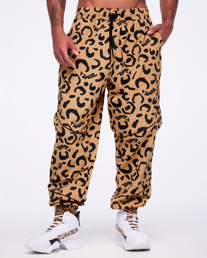 Tan leopard-print cargo pants with black abstract spots, flap side cargo pockets, a black drawstring waistband, and elastic cuffs, shown from the front on a male model wearing white high-top Zumba sneakers with leopard-print and black accents.
