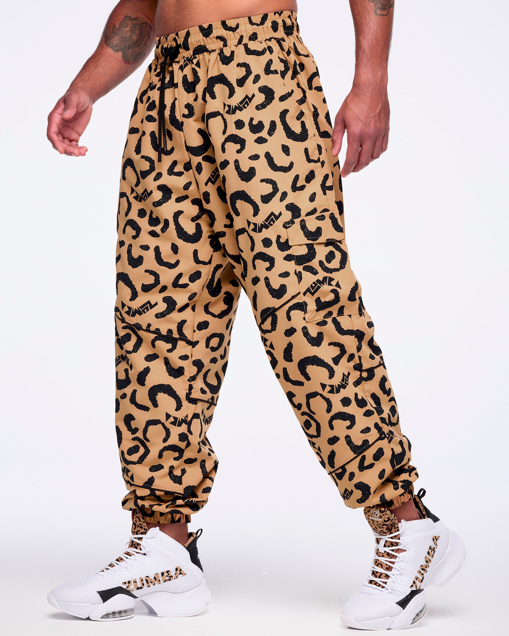 Tan leopard-print cargo pants with black abstract spots, flap side cargo pockets, a black drawstring waistband, and elastic cuffs, shown on a male model wearing white high-top Zumba sneakers with leopard-print tongues and black accents.