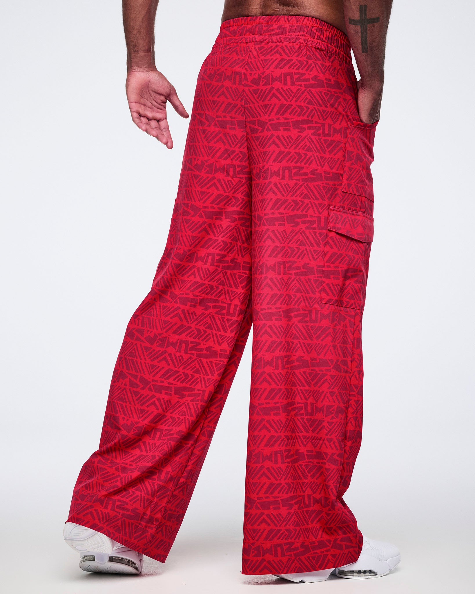 Male model wearing wide-leg red cargo pants with a tonal geometric Zumba print, shown from the back with an elastic waistband, side cargo pocket, and white athletic sneakers.