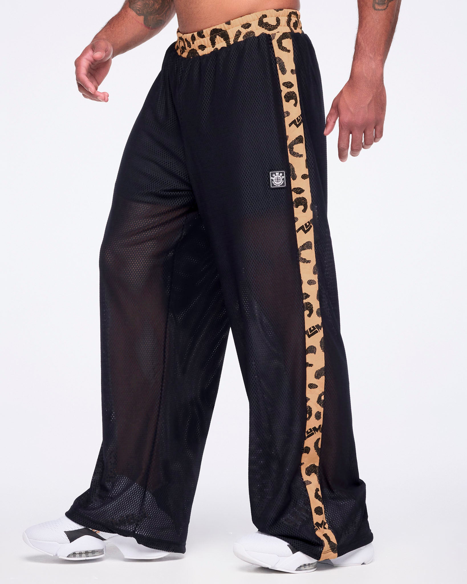 Black mesh wide-leg pants with a sheer finish, tan leopard-print waistband, matching leopard-print side stripes, and a small front logo patch, shown on a male model wearing white sneakers.
