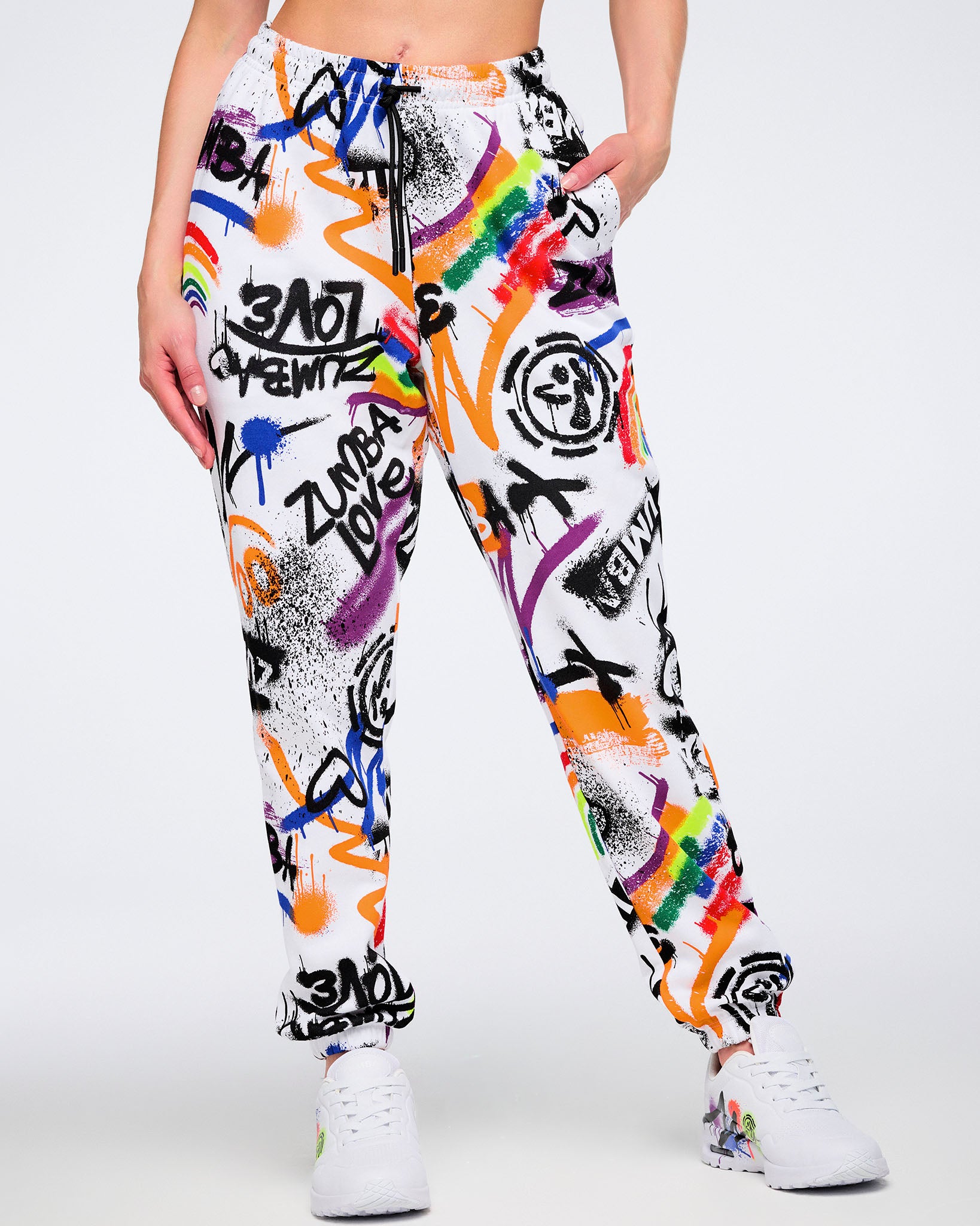 Female model wearing white and colorful graffiti-style pants with her hand in the left pocket and visible drawstring wearing matching graffiti-style shoes against a white background.