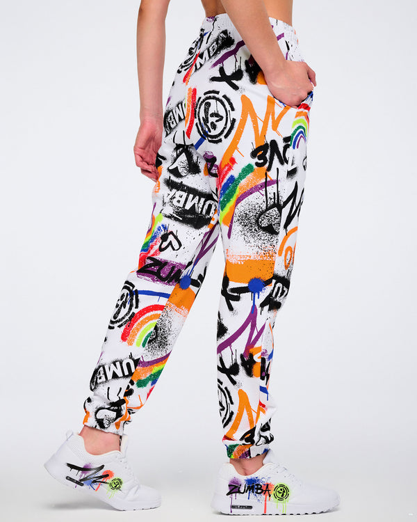 Female model shown from the back, wearing white and colorful graffiti-style pants with her hand in the right pocket wearing matching graffiti-style shoes against a white background.